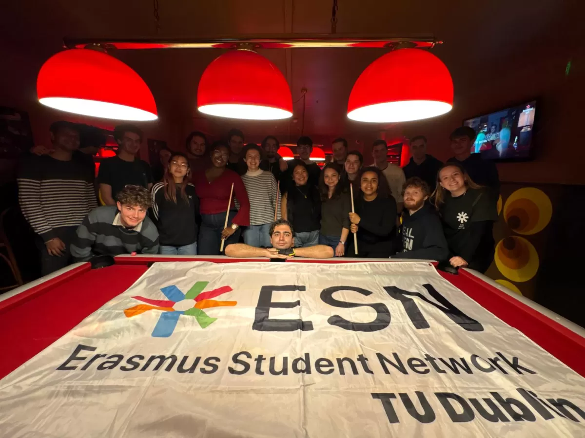 A group of students smiles and holds pool cues around a billiard table. A large "Erasmus Student Network" flag covers the table. Red lamps above.