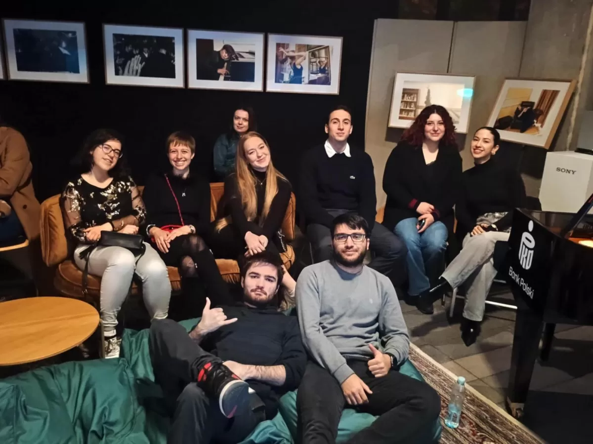 A group young adults sit and lounge together in a cozy indoor space. Five people sit on a couch and two on chairs behind a piano, while two others sit and lie on beanbags in front. They are smiling and relaxed. The background features framed photographs on a dark wall.