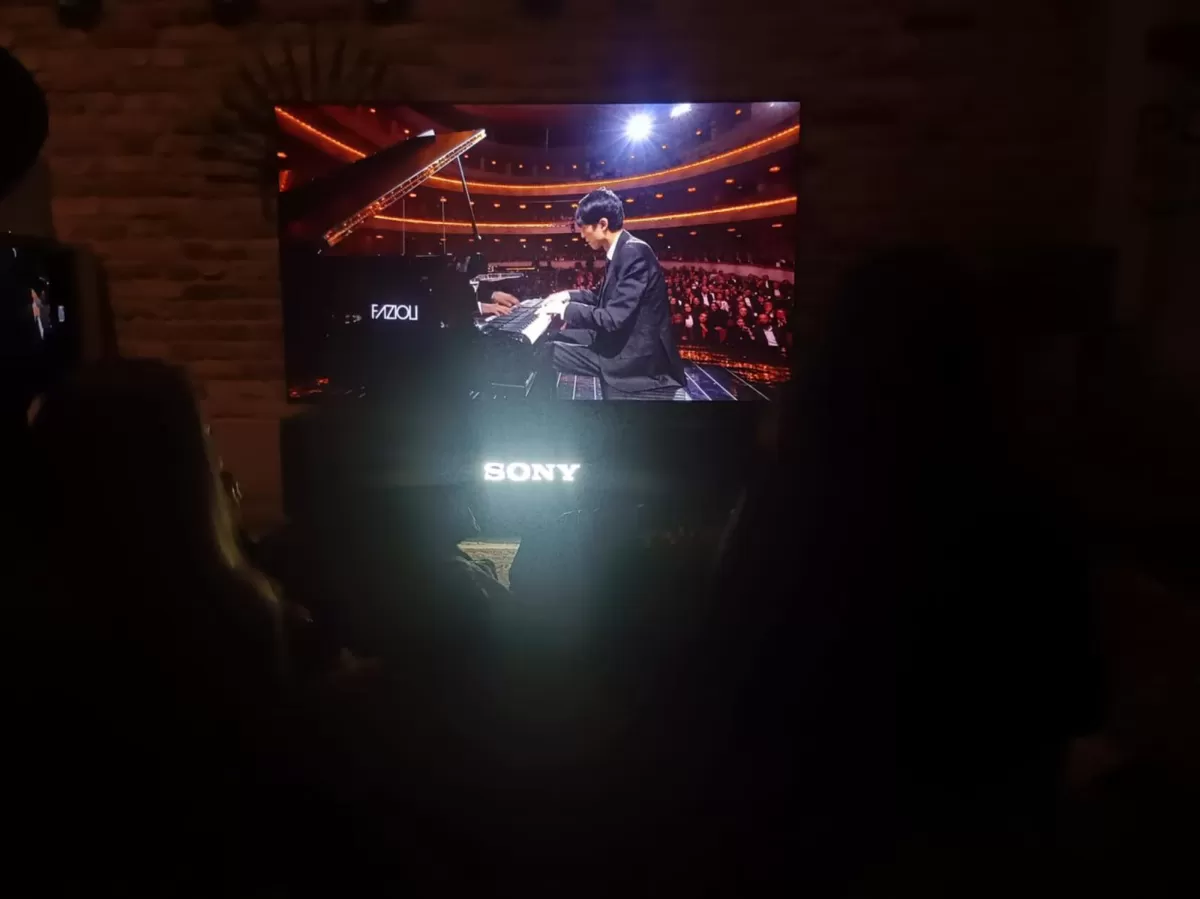A TV screen shows a pianist performing onstage in a large concert hall filled with an audience. The room where the photo is taken is dimly lit, with silhouettes of people watching the performance. The glowing “SONY” logo is visible beneath the screen.