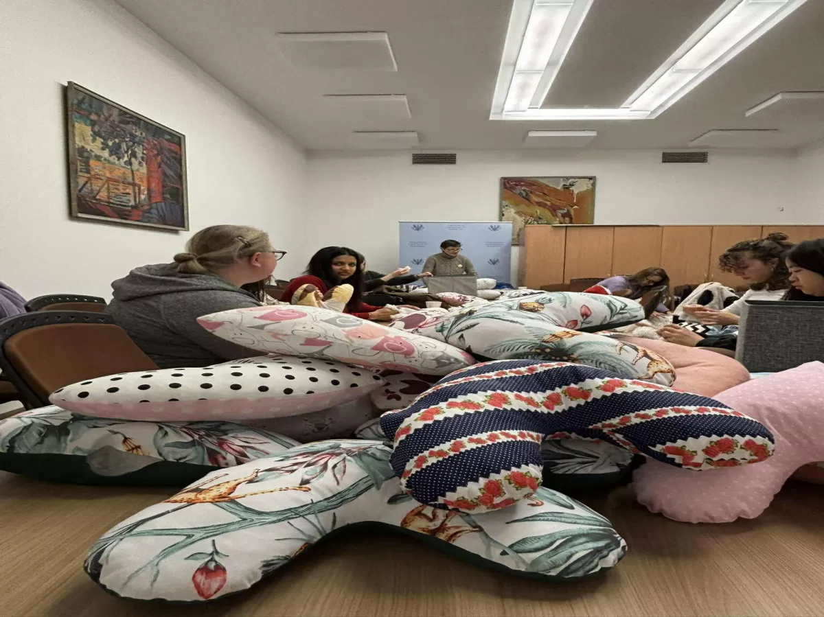 An image showing a group of several people sitting around a long table in a well-lit room, possibly for a crafting workshop or meeting. The foreground is dominated by a large pile of colorful, U-shaped and crescent-shaped pillows with various patterns, including white with black dots, floral designs with leaves and animals, and a dark blue one with red strawberries and white dots. In the middle ground, people are seated around the table, appearing to be working on similar pillows or engaged in discussion. A