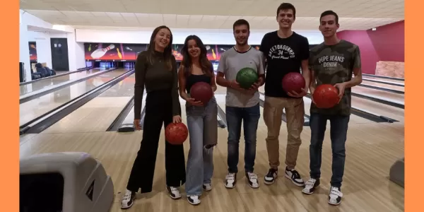 The picture shows 3 boys and 2 girls standing next to each other and holding a bowing ball, and all of them are smiling at the camera. In the background there are several bowling tracks.