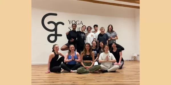 The participants are in a group looking at camera and smiling. Some of them are standing and some are sitting on the wooden floor of the yoga studio.