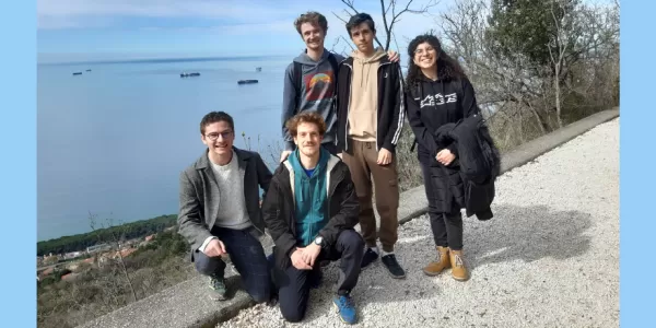The picture shows 4 boys and a girl standing together in a group on a hiking trail, looking at the camera and smiling. There is the sea in the background, as well as a few tree branches and blue sky.