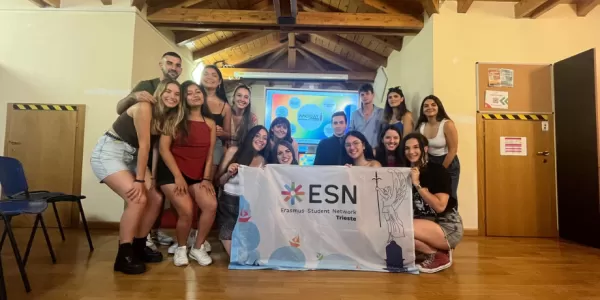 The perticipants are standing in a group smiling at the camera. They are holding ESN Trieste's flag. Behind them, a presentation is being shown which displays Arcigay's logo.