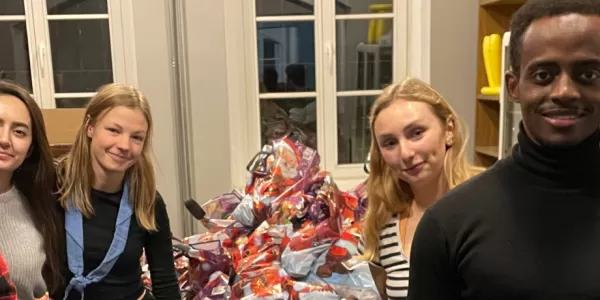 Erasmus students standing in two lines, holding the ESN Toruń flag between them. Behind them is a mountain of colorful parcels that they packed for the residents of Toruń