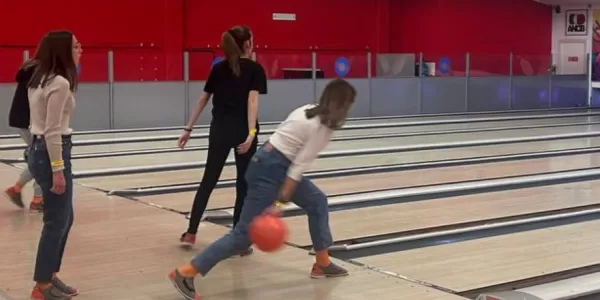 In the photo, a girl is captured in the midst of her bowling action, her focus intense as she swings her arm forward to release the ball. Her posture is dynamic, embodying both concentration and grace.