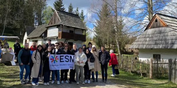 a group of students standing before a traditional houses in a museum holding a ESN UNIZA flag