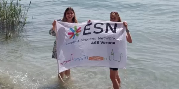 Two international students holding the ESN ASE Verona flag while standing in the lake