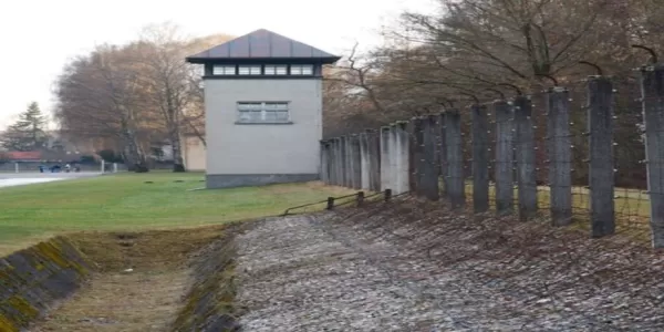 A concentration camp with a watchtower, barbed wire fence, and people walking along a pathway.