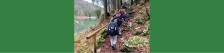 A group of people are walking on a path in the woods. On the left, a lake with very clear water is in the background.