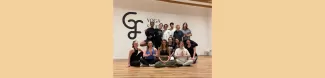 The participants are in a group looking at camera and smiling. Some of them are standing and some are sitting on the wooden floor of the yoga studio.