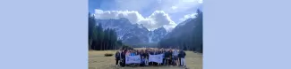 The participants are standing in a group holding the flags of the different sections. Behind them there are some trees and a mountain with snow of the top.