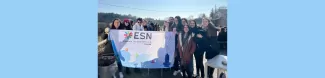 The participants are standing in front of the lunch table, holding ESN Trieste's flag and smiling. A blue sky and a green hill landscape are in the background.