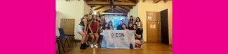 The perticipants are standing in a group smiling at the camera. They are holding ESN Trieste's flag. Behind them, a presentation is being shown which displays Arcigay's logo.