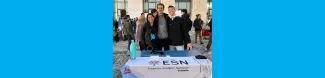 Three ESN volunteers are looking at the camera and smiling. They are standing behind the ESN table at the University's Welcome Day fair. ESN Trieste's flag is laying on top of the table.