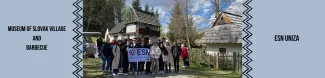 a group of students standing before a traditional houses in a museum holding a ESN UNIZA flag