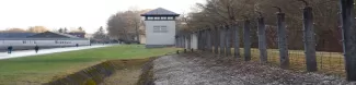 A concentration camp with a watchtower, barbed wire fence, and people walking along a pathway.