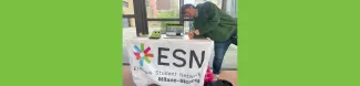 A volunteer, Cocco the crocodile, a computer showing AAC symbol website and ESN Milano-Bicocca flag