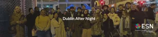 A group of people smiling at night in Dublin, dressed warmly. Bright streetlights and a red traffic light are visible. Text reads "Dublin After Night." ESN logo in the corner.